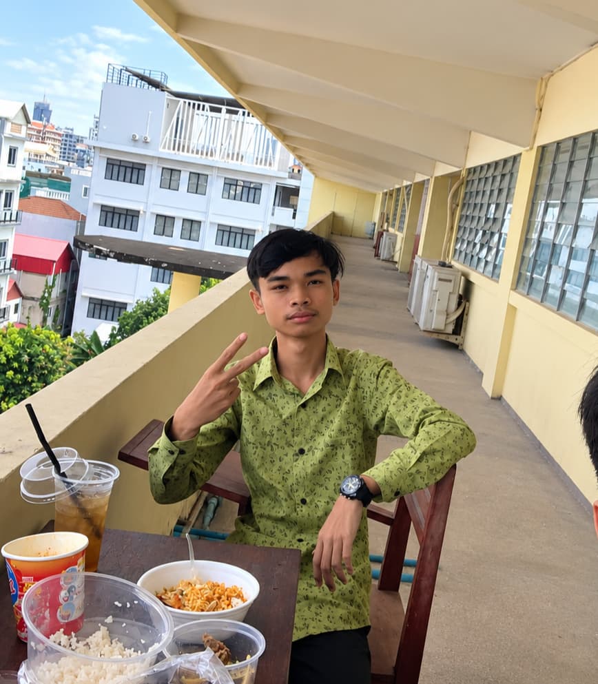 Two friends taking a selfie on a sunny open-air corridor, with a table of lunch in the foreground and Phnom Penh buildings behind them